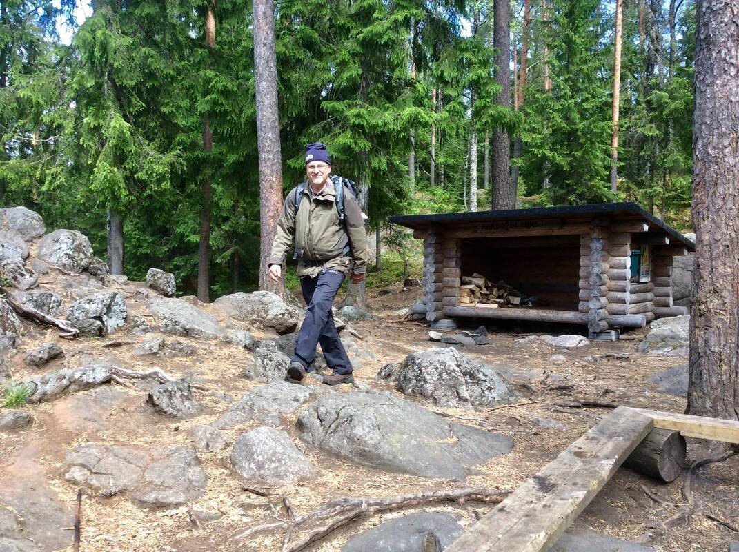 A guide walking past a log lean-to shelter among pines and boulders in the Usmi wilderness forest