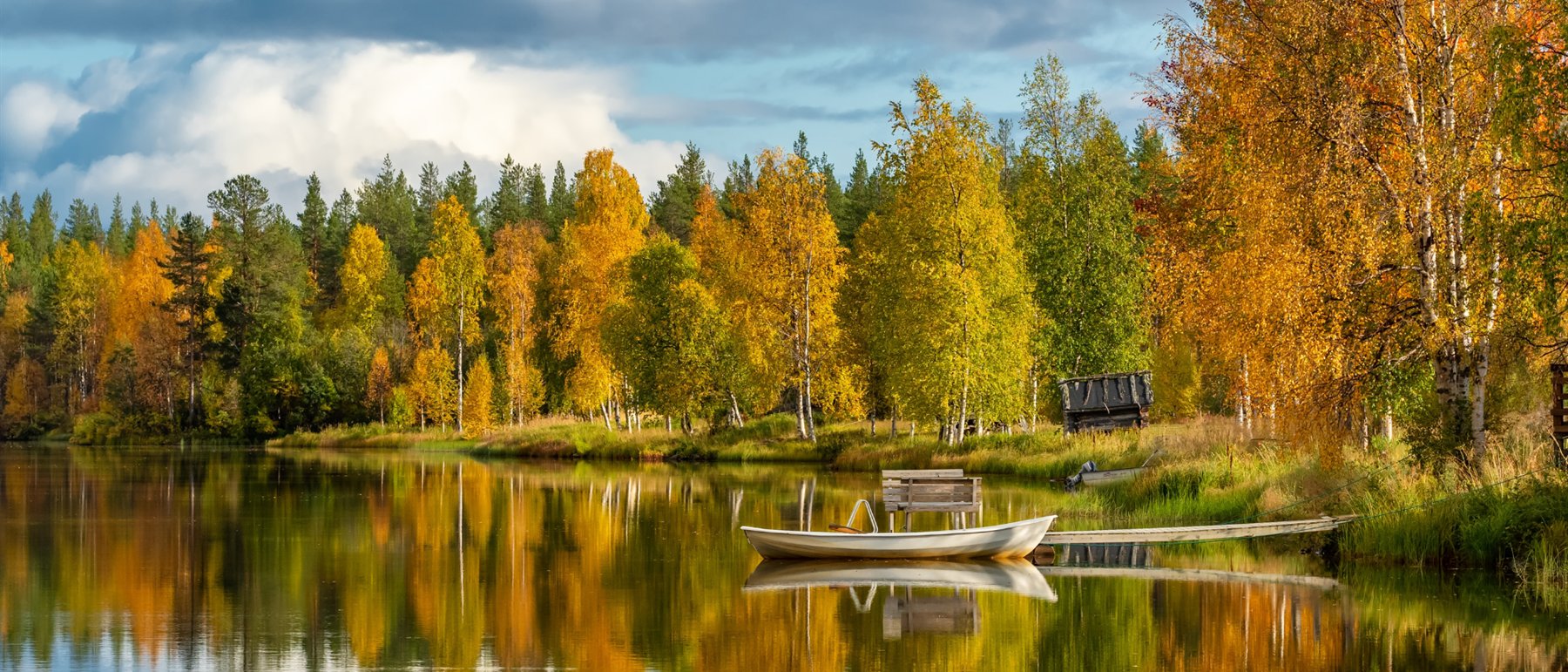 A still Finnish lake in autumn. Birch and aspen along the far shore have turned gold and orange against dark spruce, a white rowing boat is moored by a small wooden jetty, and the shoreline is mirrored in the flat water under a cloudy blue sky.